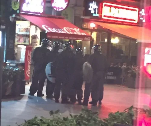 Riot police in protective gear stand in formation outside lit storefronts in Leicester Square at night.