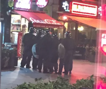 Riot police in protective gear stand in formation outside lit storefronts in Leicester Square at night.