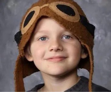 A child wearing a brown aviator hat with floppy ears, smiling slightly against a gray background.