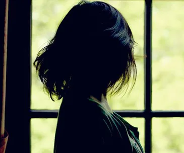 A woman with short dark hair stands in shadow facing a window with light streaming in.