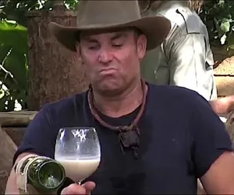 Man wearing a hat making a funny face while pouring a drink into a glass, outdoors in a jungle camp setting.