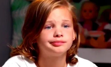 A young child with shoulder-length hair looks emotional, sitting indoors with dolls in the background.