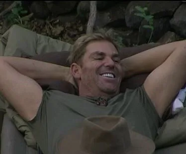 Man relaxing outdoors with hands behind head, smiling broadly, wearing a green shirt; a hat lies in the foreground.