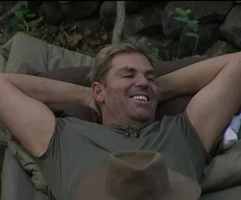 Man relaxing outdoors with hands behind head, smiling broadly, wearing a green shirt; a hat lies in the foreground.