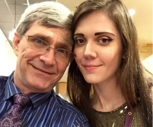 Couple smiling together, man with glasses and a patterned tie, woman with long brown hair and winged eyeliner.