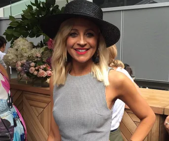 A woman wearing a black hat and a checked dress is smiling outside near flowers and a wooden table.