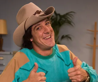 Man in beige cowboy hat and colorful sweater giving double thumbs up, smiling indoors.