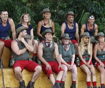 Group of people in jungle-themed outfits on "I'm a Celebrity" set, wearing hats and sitting on steps.