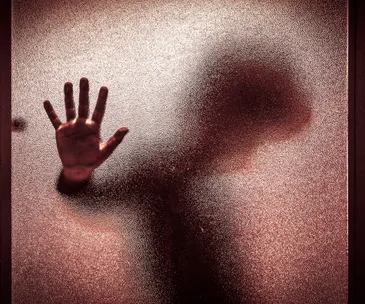 Shadowy figure with hand pressed against frosted glass, conveying a sense of confinement or distress.