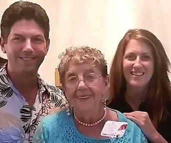 Three people smiling, with the elderly woman in the center wearing glasses and a blue top.
