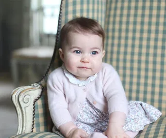 A young child, sitting in a checkered chair, wearing a pink sweater and floral dress, looks playfully to the side.