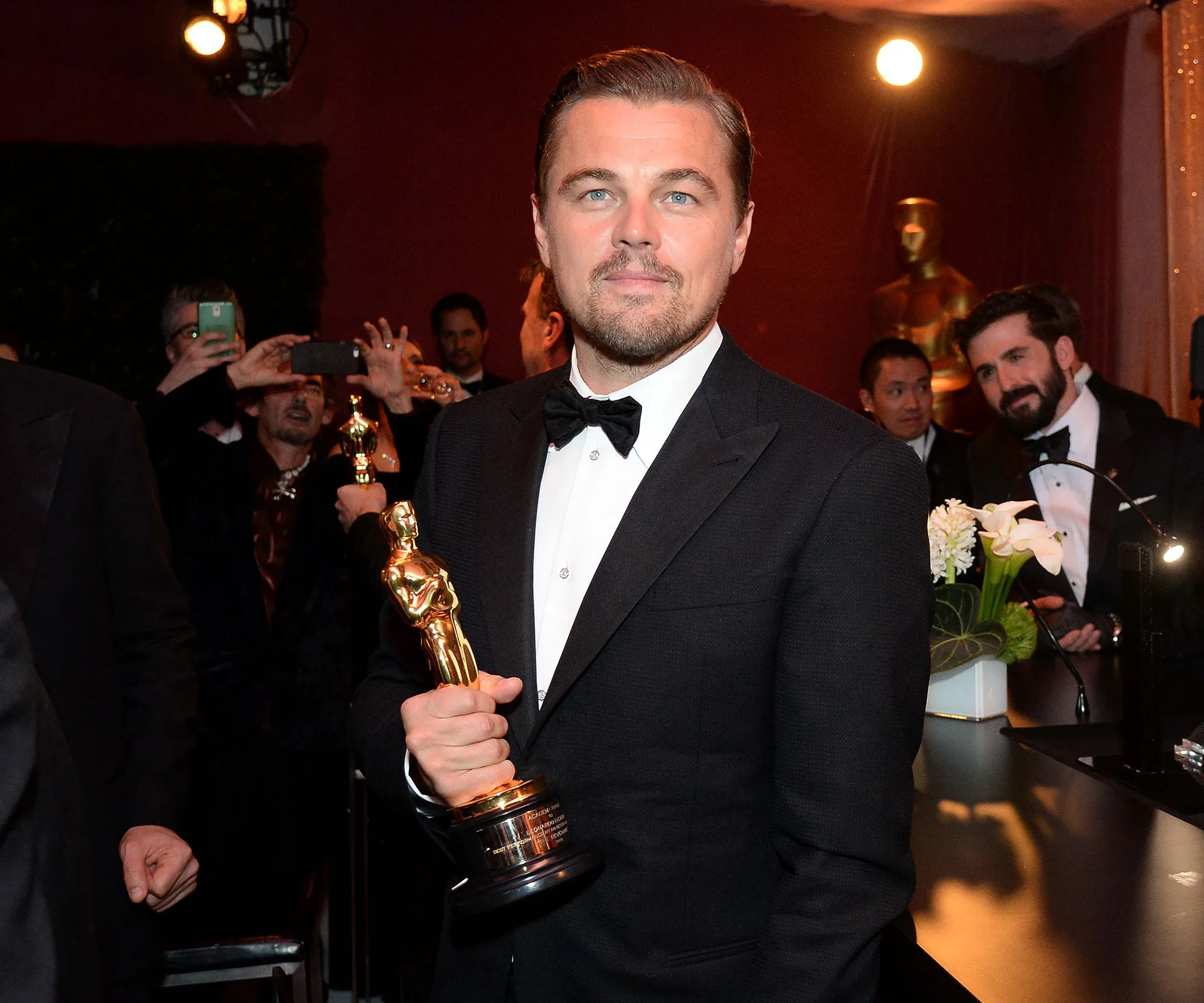 A man in a tuxedo holding an Oscar statuette, with people and cameras in the background at an event.