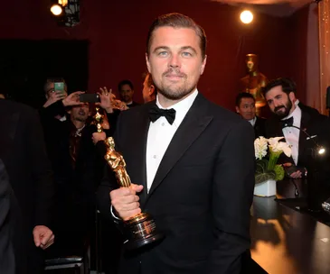 A man in a tuxedo holding an Oscar statuette, with people and cameras in the background at an event.