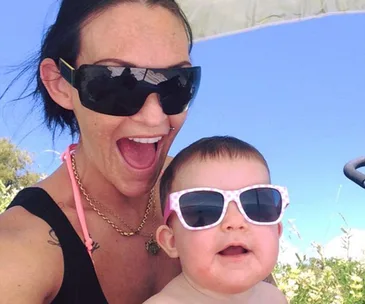 A woman and baby wearing sunglasses smiling outdoors under a clear blue sky.