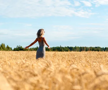 Woman in a gray dress stands joyfully in a golden wheat field under a clear blue sky.