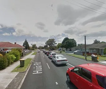 A suburban street with lined cars on Huia Road, houses, and cloudy sky.