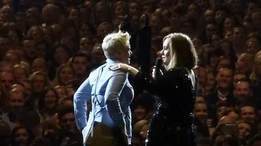 Adele on stage helps a fan propose at a concert, surrounded by a cheering crowd.