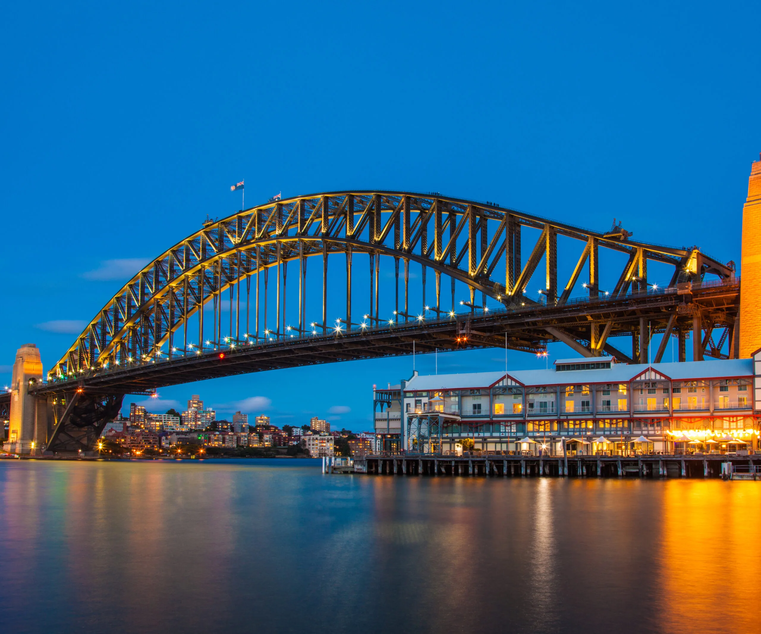 Sydney Harbour Bridge at dusk with illuminated arch, water reflections, and cityscape in the background.