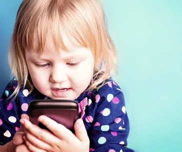A young child with blond hair, in a polka dot shirt, looks intently at a handheld device against a blue background.