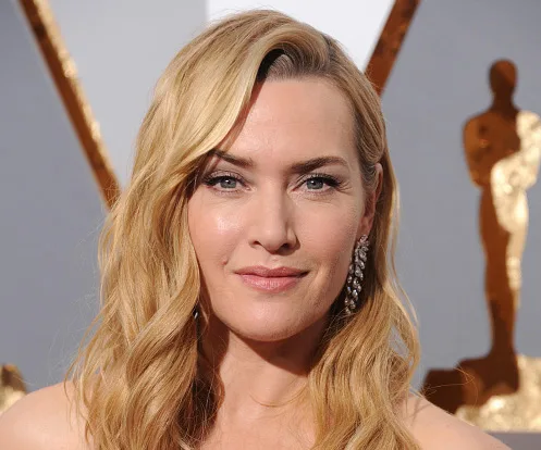 Kate Winslet at an event, wearing a strapless dress, with wavy blonde hair and statement earrings.