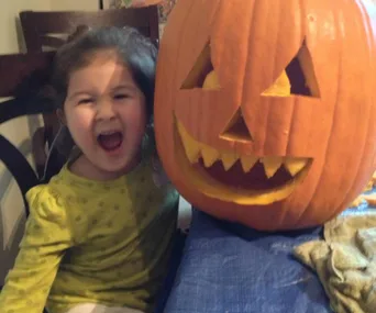 A child smiling beside a carved Halloween pumpkin with a toothy grin.