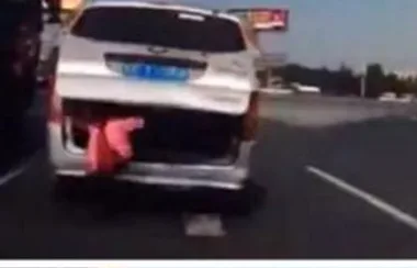 A moving car with an open trunk on a highway, a pink object hanging out, suggesting possible danger.