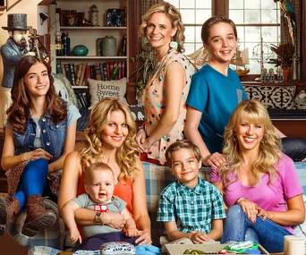 Cast of "Fuller House" smiles in a living room setting, featuring adults and children on a couch.