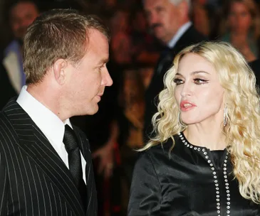 A man in a black pinstripe suit and a woman with long blonde hair and a black dress, facing each other in conversation.