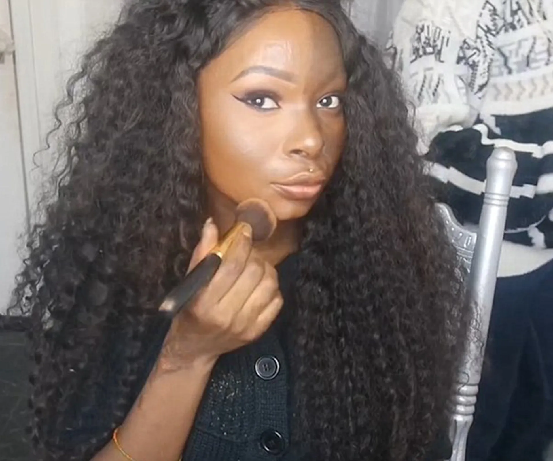A woman with long curly hair applies makeup with a brush, seated on a silver chair indoors.