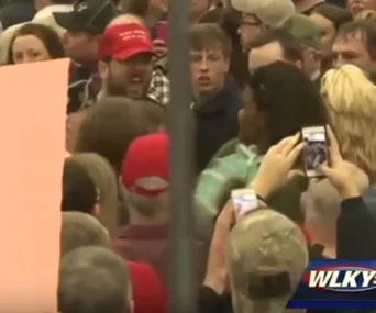 A crowd at a rally, with people capturing the event on phones. A woman is surrounded by attendees.