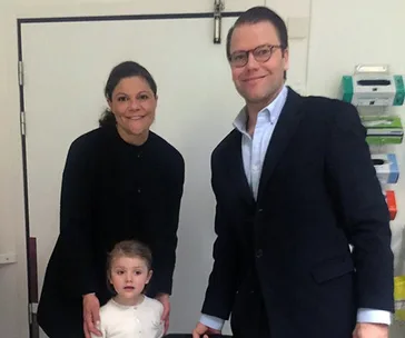 Family smiling together, with a standing child, inside a room with medical supplies in the background.