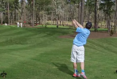 Young golfer in blue shirt swings club on lush course; trees in background.