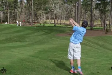 Young golfer in blue shirt swings club on lush course; trees in background.