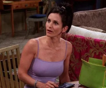 Monica sitting in her apartment on "Friends" in a purple top, with a green bag on the table beside her.