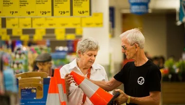 Two people examining traffic cones inside a supermarket with product signs and shelves in the background.
