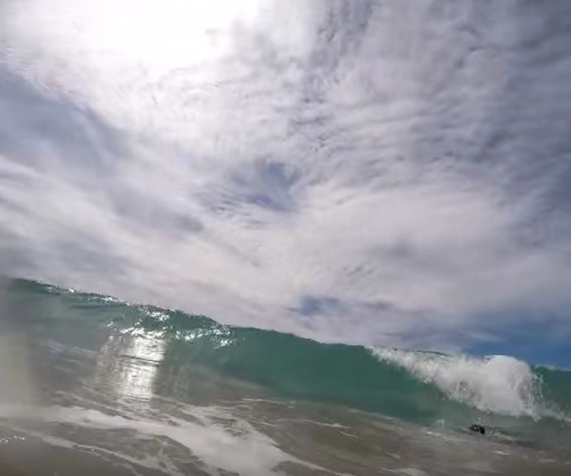 Ocean wave under a cloudy sky, with sunlight reflecting on the water's surface.