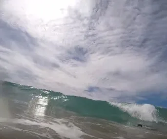 Ocean wave under a cloudy sky, with sunlight reflecting on the water's surface.