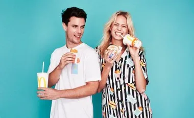 Two people smiling, holding McDonald's food and drinks, with a turquoise background; one wears a shirt with McDonald's icons.