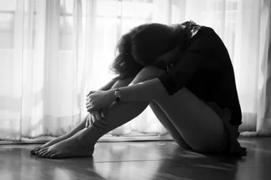 A woman sitting on the floor with her head resting on her knees in a dimly lit room.