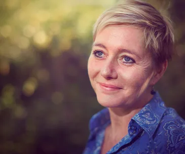Smiling woman with short blonde hair in a blue patterned shirt, outdoor background.