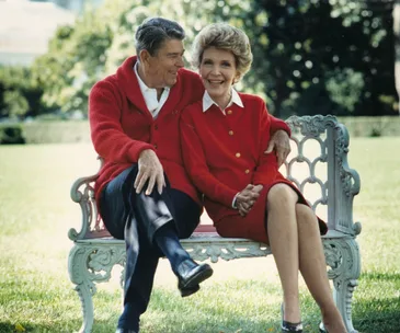 A couple in red clothing sitting on a bench, smiling in an outdoor garden setting.