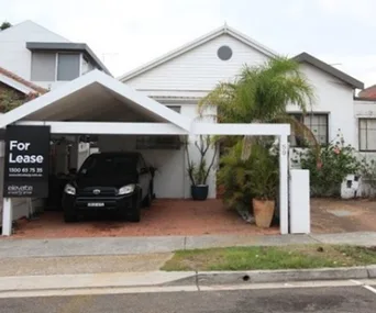 House with driveway and car under a "For Lease" sign, surrounded by plants and adjacent homes.