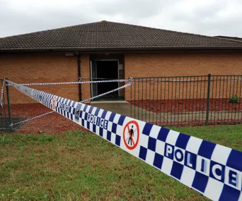 Police tape outside a brick building with grass in the foreground, indicating a restricted area.