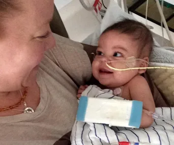 A smiling baby with a nasal tube in a hospital bed looks at an adult.