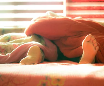 Feet peeking out from under vibrant orange and pink blankets in a bright, sunlit room.