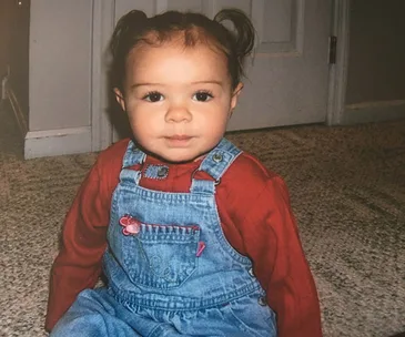 A toddler with pigtails wearing a red shirt and denim overalls sits on a carpeted floor, looking at the camera.