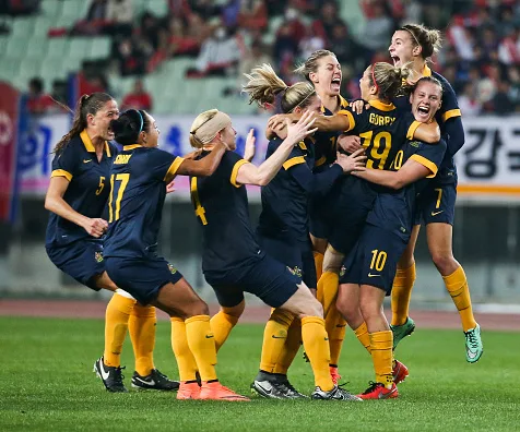 Australian women's soccer team jubilantly celebrating on the field after qualifying for the Olympics, wearing blue and yellow.