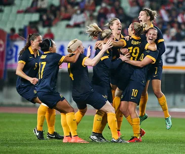 Australian women's soccer team jubilantly celebrating on the field after qualifying for the Olympics, wearing blue and yellow.