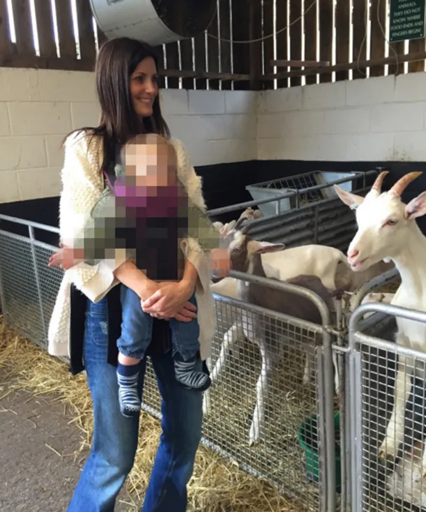 A woman holds a pixelated child near a goat pen in a barn setting.