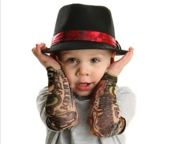 Toddler wearing a black fedora with a red band, showing off tattoo sleeves on both arms, posing with hands on cheeks.